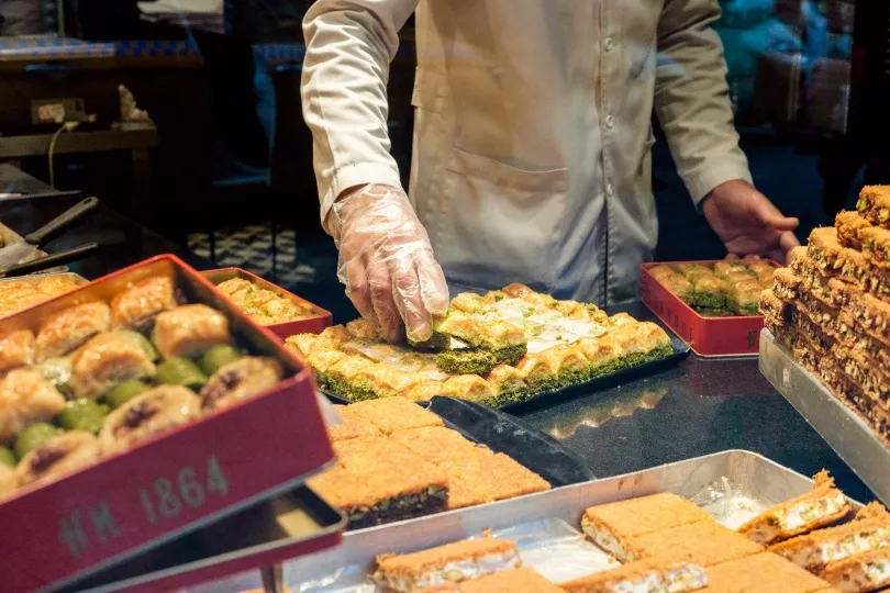 Marktkraam in Griekenland met allerlei soorten gebakjes en baklava en een verkoper die baklava pakt.