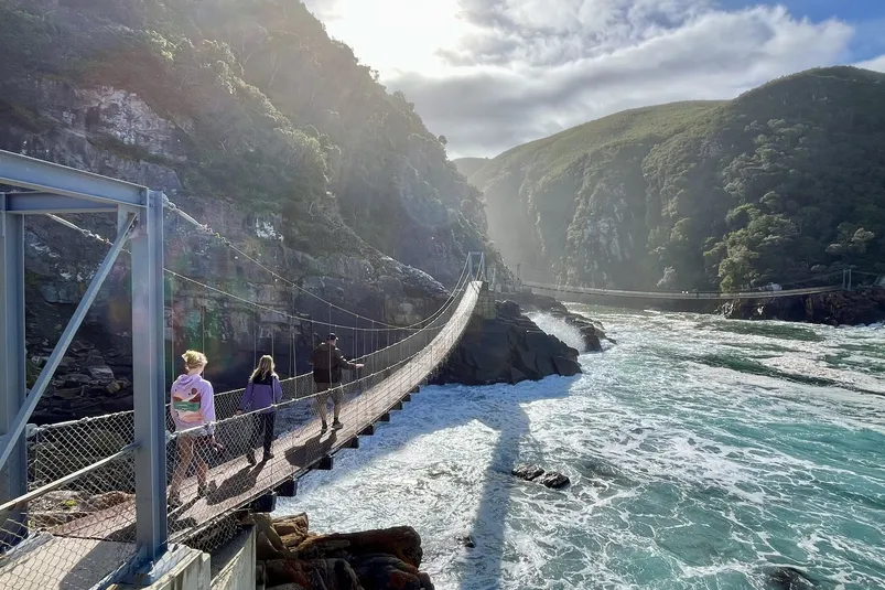Je ziet drie mensen die over een hangbrug lopen boven ruige golven, omringd door steile groene kliffen en zonlicht dat door de wolken breekt.
