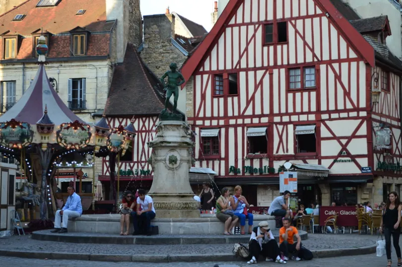 Een plein in Dijon waar mensen zitten voor een fontein met een standbeeld, omringt door terrasjes en een draaimolen met omliggende vakwerkhuizen uit de middeleeuwen en een architectuur met strepen over de huizen.