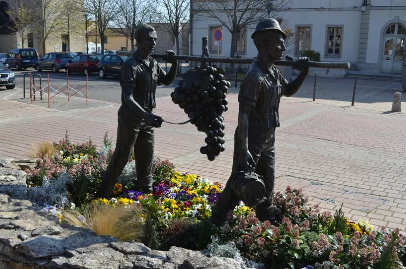 Standbeeld van twee mannen op een pleintje die een stop op hun schouders dragen met daaraan een hangende tros druiven en de voorste man heeft in zijn rechter hand nog een wijnton. Ze staan in een perkje van bloemen wat afgebakend is met stenen. 