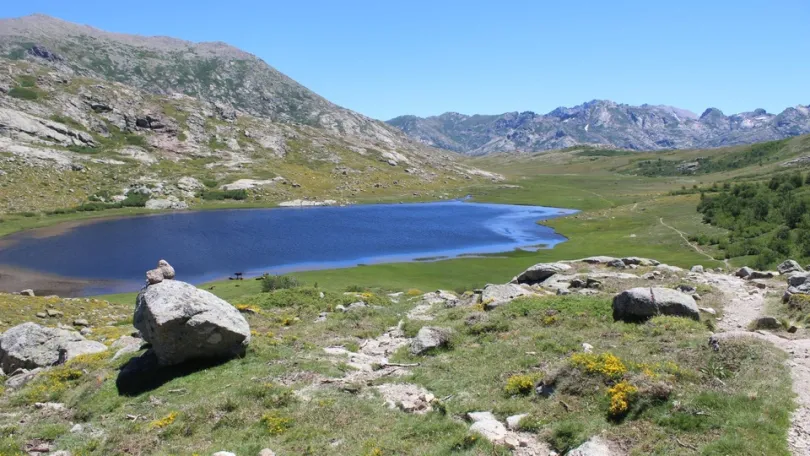 Uitzicht tussen twee bergtoppen waar het meer de Nino ligt in Corsica met groene velden en in de verte meer bergtoppen.