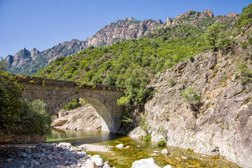 Een oude Genovese stenen brug over de Tavulella-stroom in de Spelunca-kloof met turkoois helder water in het zonlicht en de blauwe lucht.
