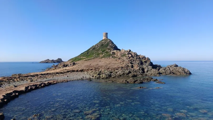 In Corsica bij het plaatjes Parata uitzicht op een rustige, rotsachtige kust steekt als een smalle heuvel uit de helderblauwe zee, met bovenop een oude stenen toren onder een wolkeloze lucht die een gevoel van stilte en ruimte uitstraalt.