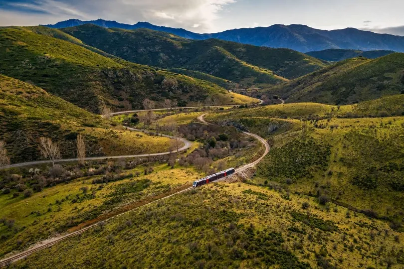 Een trein rijdt door een uitgestrekt berglandschap met glooiende heuvels en kronkelende wegen, omringd door rustige natuur en hoge bergketens op de achtergrond.