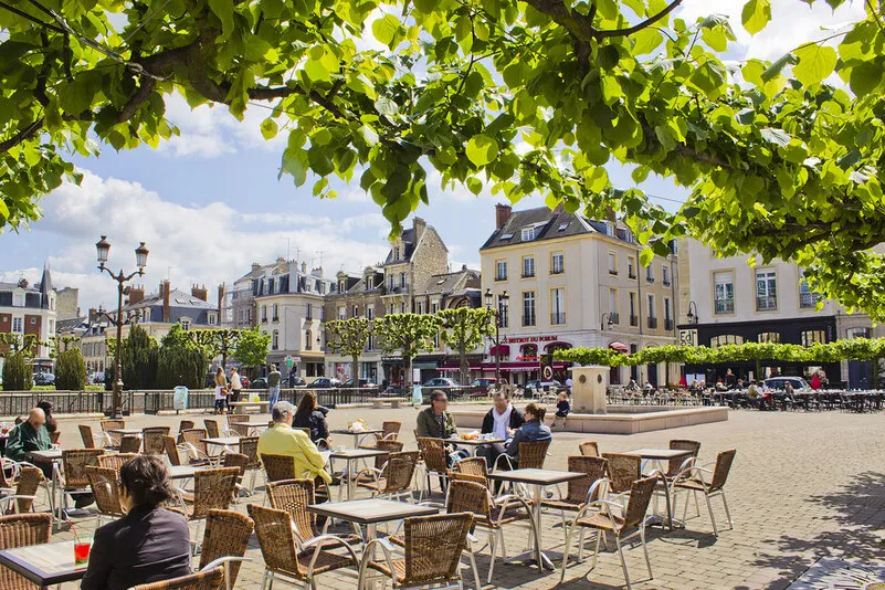 Een gezellig plein met terrassen en bezoekers in Reims dat wordt omringd door historische gebouwen en groene bomen, die voor een sfeervolle en levendige uitstraling zorgen.