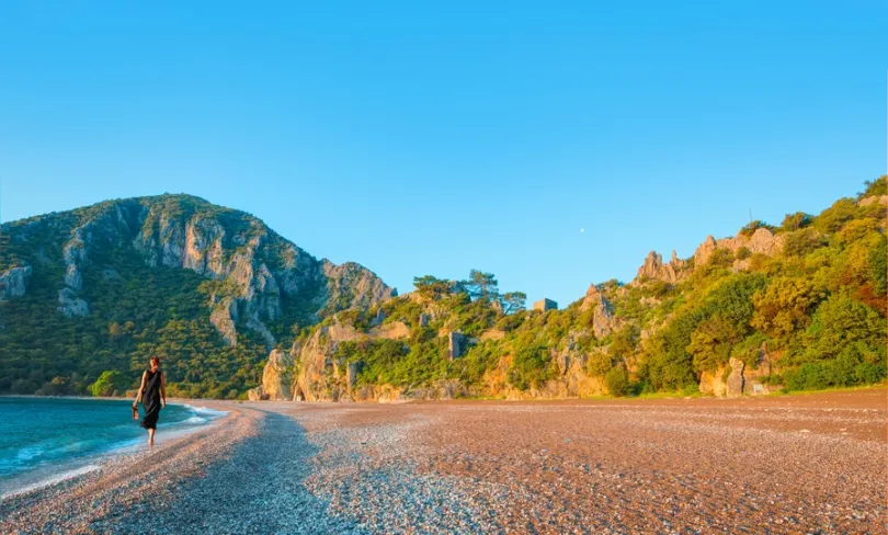 Vrouw die loopt over het rustige strand Çıralı met groene bergen op de achtergrond.