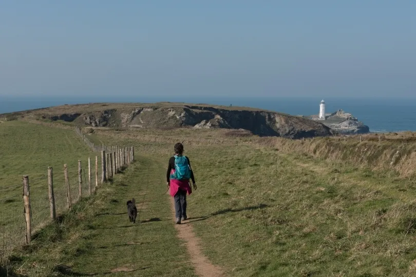 Een wandelaar loopt met een hond over een kustpad richting een vuurtoren op de kliffen.