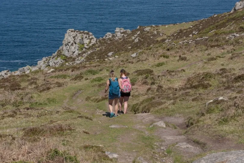 Twee wandelaars lopen over een smal kustpad door een heuvelachtig landschap met uitzicht op zee.