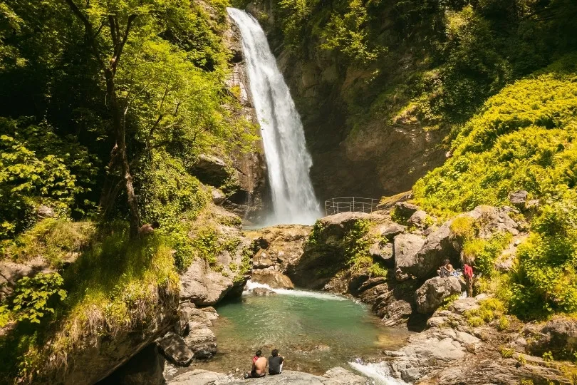 Een hoge waterval die uitkomt in een helder natuurbad, omringd door rotsen en groen.