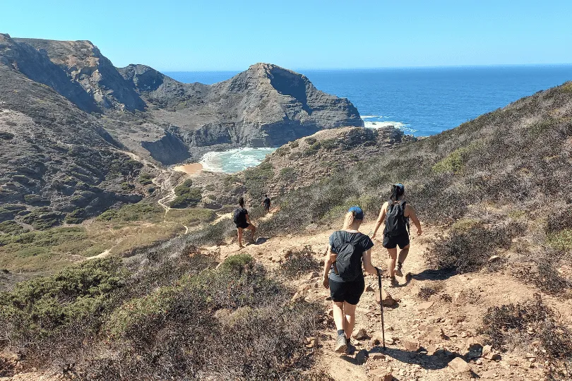 Wandelaars op een zandpad langs de kust met uitzicht op groene heuvels.