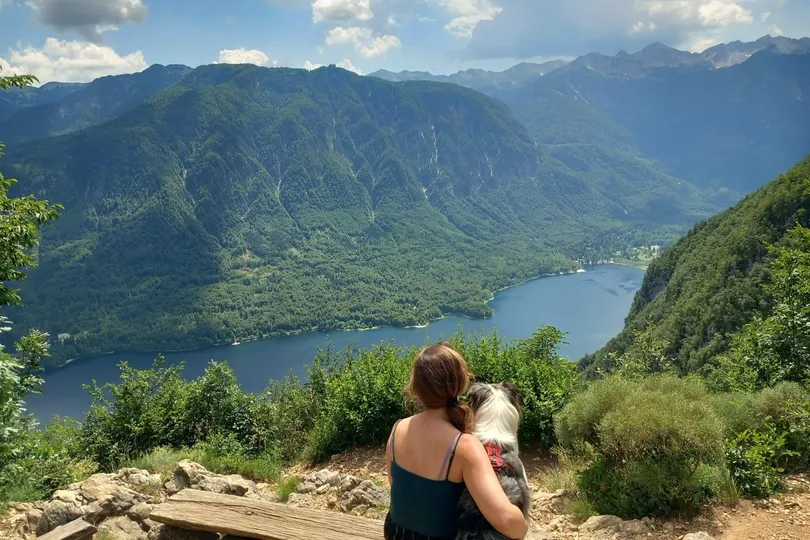 Vrouw met hond op bankje uitkijkend op meer en bergen.
