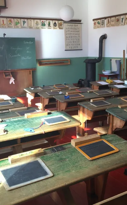 Primitief klaslokaal met schrijfbordjes op houten bankjes en een ouderwets schoolbord aan de muur.