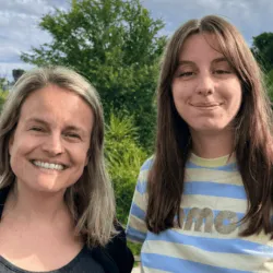 Twee lachende vrouwen naast elkaar met een groene struiken en bomen en een blauwe lucht achter hen.