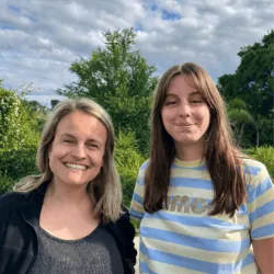 Twee vrouwen die lachend poseren voor wat groene bossen met een blauwe lucht met wat wolken.