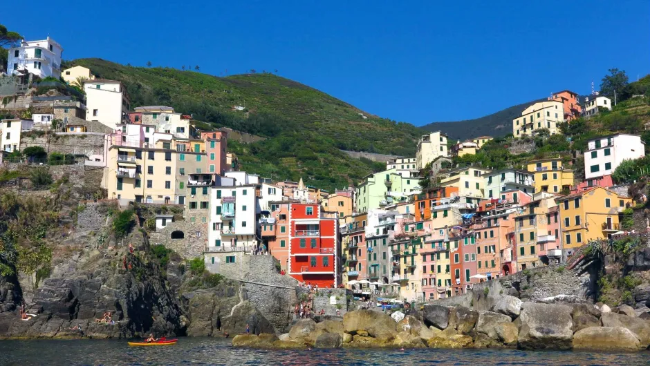 Kleurrijk kustdorp in Cinque Terre met pastelkleurige huizen tegen een groene heuvel, rotsen aan zee en een kajak op het heldere water.