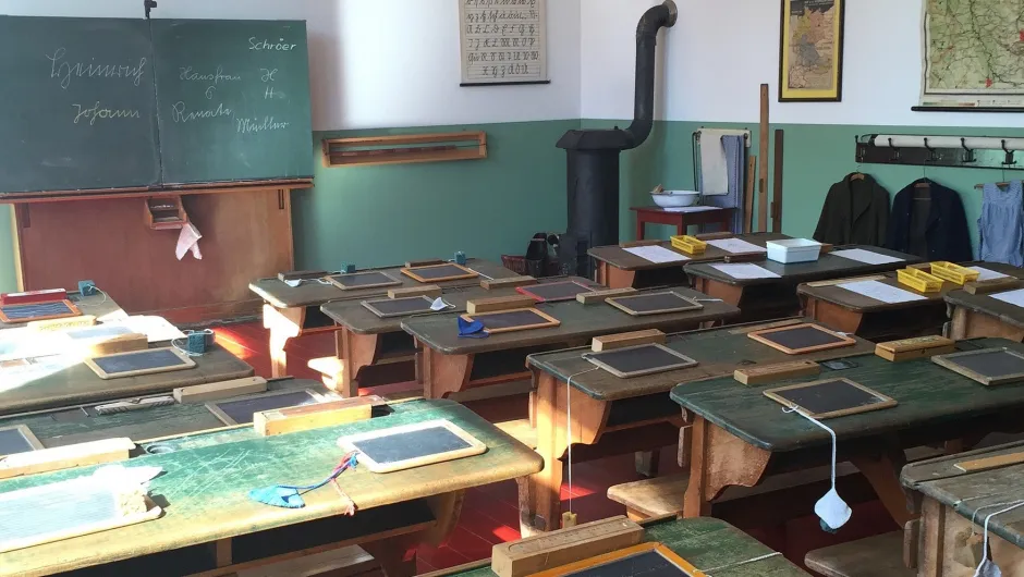 Primitief klaslokaal met schrijfbordjes op houten bankjes en een ouderwets schoolbord aan de muur.