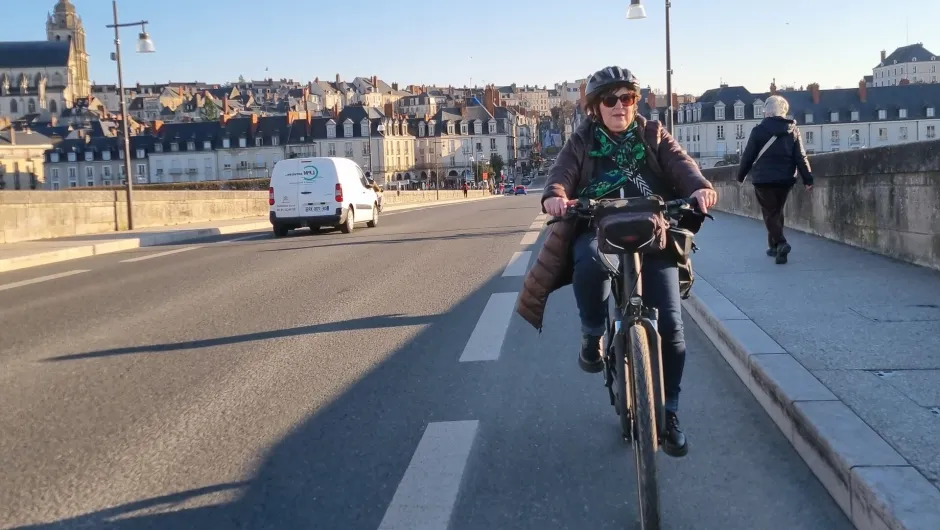 Vrouw die aan het fietsen is over een brug met wat auto's op de achtergrond en mensen die aan de zijkant lopen. Ze heeft een help op en haar jas is open en wappert door de wint.