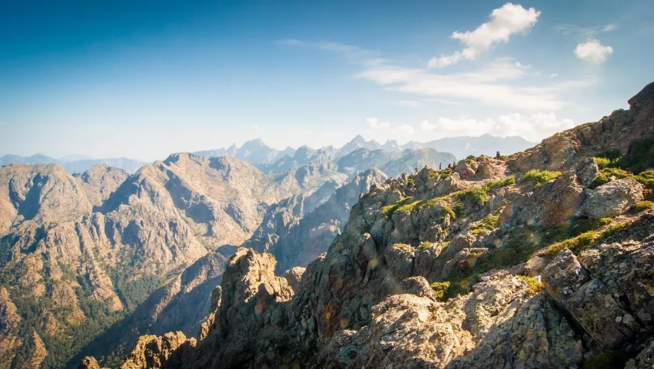 Een uitgestrekt berglandschap met ruige rotsen en hoge bergpieken strekt zich uit onder een heldere lucht, terwijl een smal wandelpad langs steile berghellingen loopt.