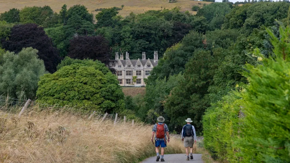 Twee wandelaars lopen door een groen landschap richting een statig landhuis tussen de bomen.