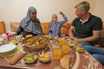 Een witte vader en dochter die met een Marokkaanse mevrouw in haar huis aan het eten zijn. 