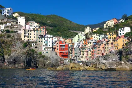 Kleurrijk kustdorp in Cinque Terre met pastelkleurige huizen tegen een groene heuvel, rotsen aan zee en een kajak op het heldere water.