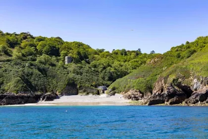 Een rustig strand met helder blauw water, omgeven door groene heuvels en een oude wachttoren bovenaan.
