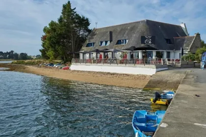 kleinschalig hotel gelegen aan de waterkant met een terras dat uitkijk op het water met daaronder een standje en bootjes die aan de zijkant vast liggen.