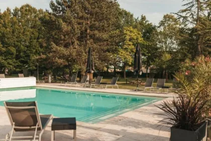 Buitenzwembad bij een hotel in een groene tuin met bomen en planten in plantenbakken met meerder ligbedjes en parasols.