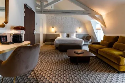 Een ruime en sfeervol ingerichte hotelkamer met een comfortabel bed, een zithoek en zichtbare houten balken onder een schuin dak straalt warmte en rust uit.