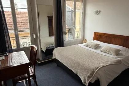 Een lichte hotelkamer met een tweepersoonsbed, houten meubels en grote ramen die uitzicht bieden op de omliggende bebouwing.