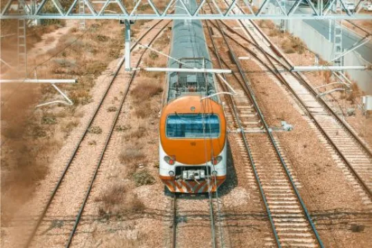 Een oranje trein die over een zonnig spoorlandschap rijdt terwijl je van bovenaf meekijkt.