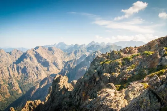Een uitgestrekt berglandschap met ruige rotsen en hoge bergpieken strekt zich uit onder een heldere lucht, terwijl een smal wandelpad langs steile berghellingen loopt.