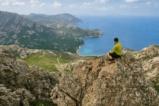 Mannelijke wandelaar die op een rots zit en uitkijkt naar de kustlijn en de bergen van Corsica.