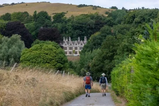Twee wandelaars lopen door een groen landschap richting een statig landhuis tussen de bomen.
